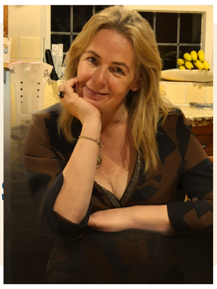 A woman smiling, wrapped in a large grey blanket, sitting at a table in a café.