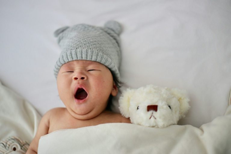 Baby wearing a grey hat yawning, lying beside a toy bear on a soft blanket.